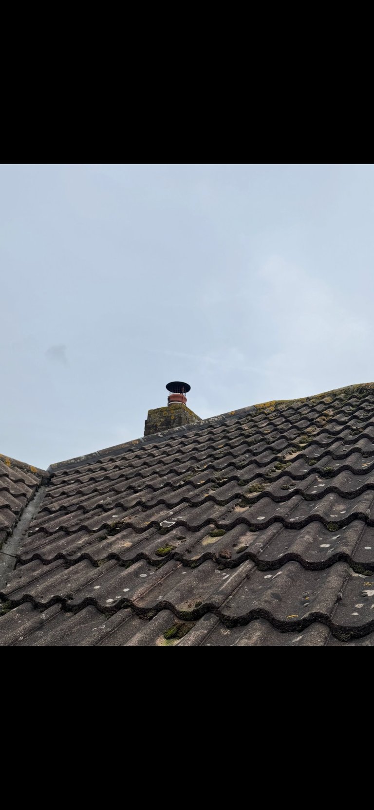 Close-up of weathered dark tile roof with chimney and moss growth against cloudy sky