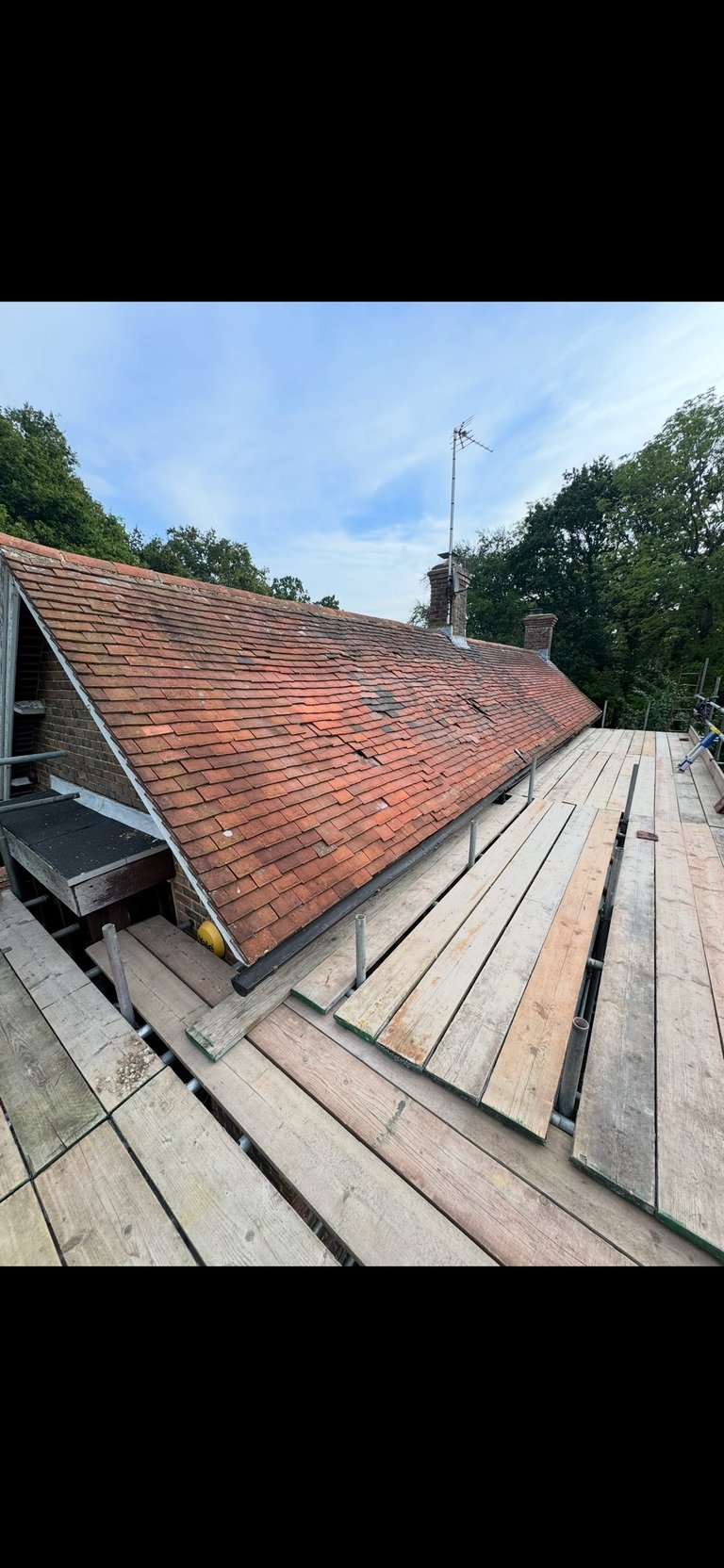 Residential roofline with terracotta clay tiles, wooden deck, and chimney stacks against blue sky with surrounding trees