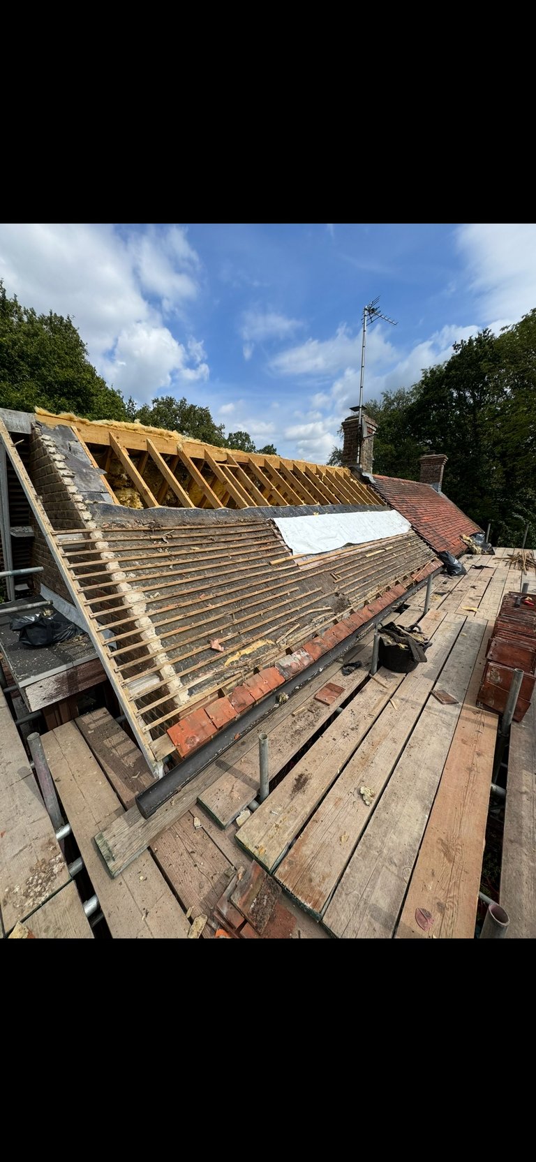 Roof under construction showing wooden framework, exposed rafters, brick chimney, and blue sky with clouds