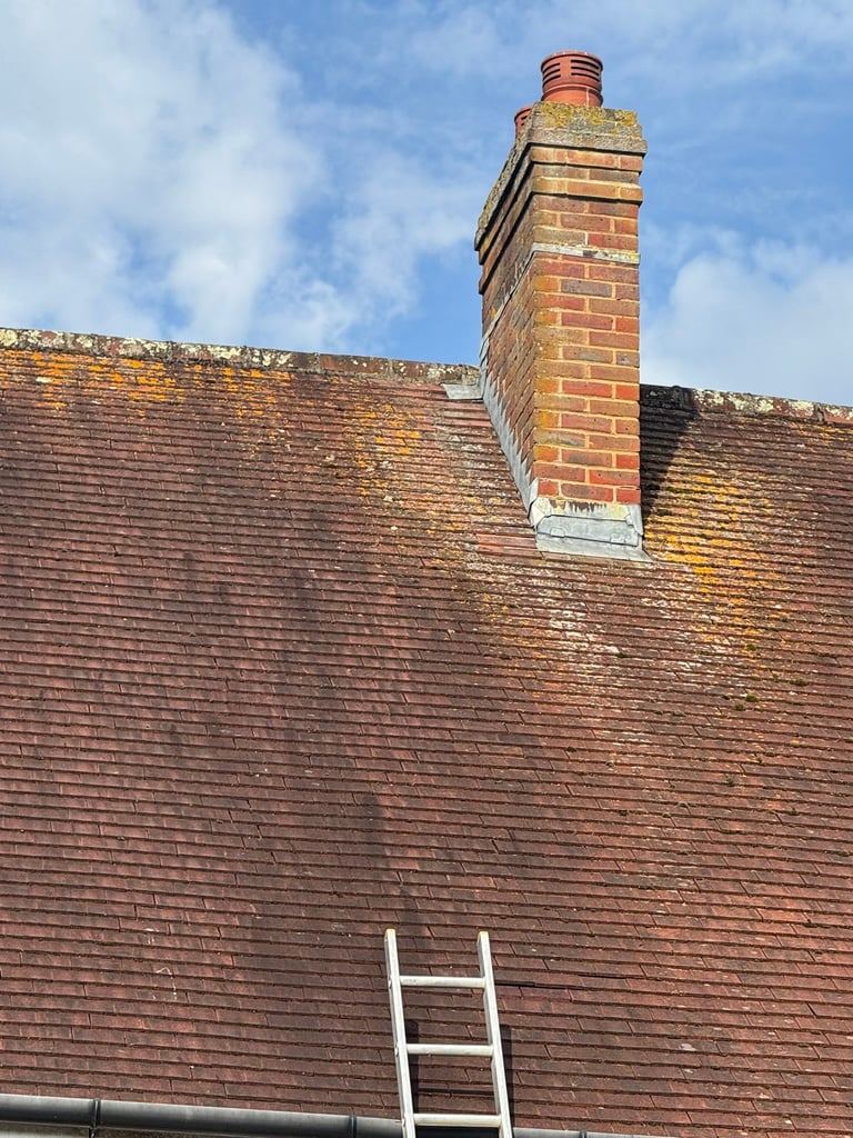 Brick chimney protruding from a red-tiled roof against a blue sky, with a white ladder leaning against the roof below.