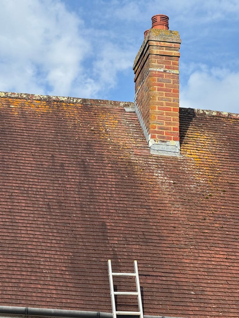 Brick chimney protruding from red clay tile roof with white ladder leaning against tiles and blue sky above, showing the repaired slipped tile