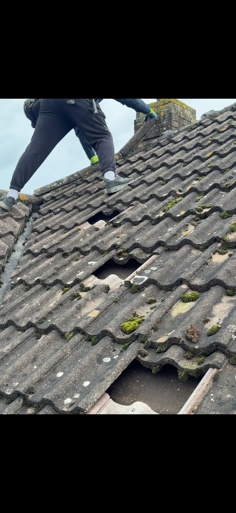 Person in dark pants and neon yellow socks climbing a steep dark roof with moss and damaged tiles showing holes